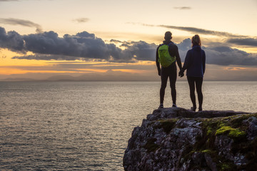 Adventurous couple holding hands and standing on a rocky cliff by the Pacific Ocean during colorful cloudy sunset. Taken in Lighthouse Park, West Vancouver, BC, Canada.