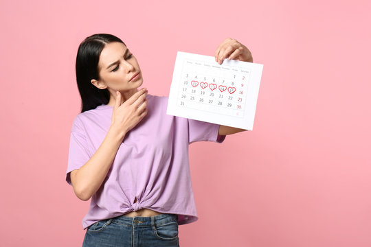 Pensive young woman holding calendar with marked menstrual cycle days on pink background