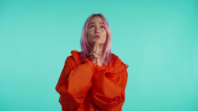 Attractive unusual woman with pink dyed hair praying and crosses her fingers over blue background. Mixed race girl begging someone.