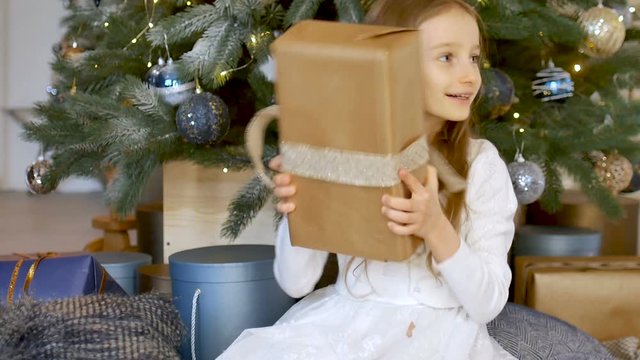 Cute blond girl has recieved a gift box wrapped in craft paper sitting near christmas tree, happy childhood and xmas celebration concepts