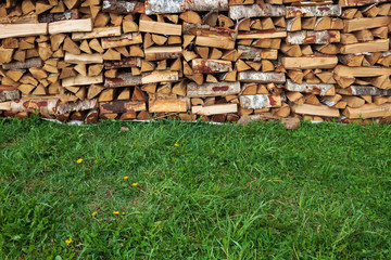 A wall of a stacked supply of firewood stands on a site covered with green grass. Background. Texture.