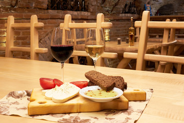 Still life with two glasses with red and white wine, brown bread, tomatoes, feta cheese and olive oil on wooden table in the old vintage wine cellar from red brick
