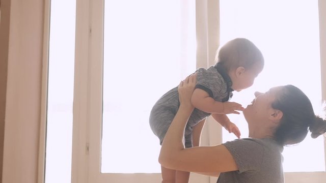 Mother and her baby son having fun and playing at home. Little kid 1 years old play with his mom arms at home near a big window, slow motion. They feel happy, smile and laugh