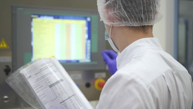 Man employee work with document and computer in interior of pharmaceutical plant. American male worker holding paper in hand and watching pc screen while working in pharma medical company. Concept