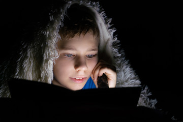 Portrait of a kid reading a book in the dark, lying in bed under a blanket