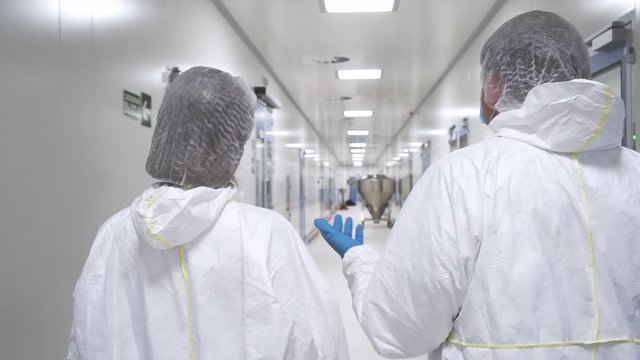 Quality Control Check: two scientists talking and wearing protective suit walking through factory hallway. Product Manufacturing. pharmacist work discussion