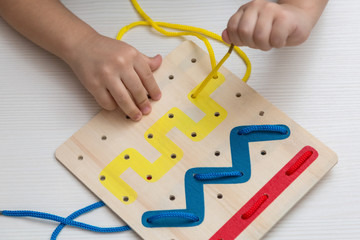 Child plays educational game interestedly with wooden colorful board and laces. Development, education.