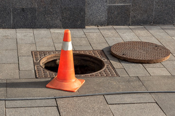 Open sewer manhole cover and traffic cone on a city street. Sewerage repair. Emergency service and accident. Drainage system maintenance. Danger sign