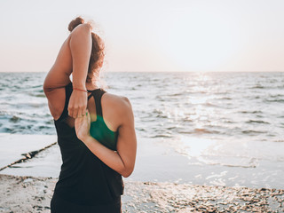 Slim woman in black bodysuit practicing yoga near sea or ocean during sunrise light. Flexibility, stretching, fitness, healthy lifestyle. 
