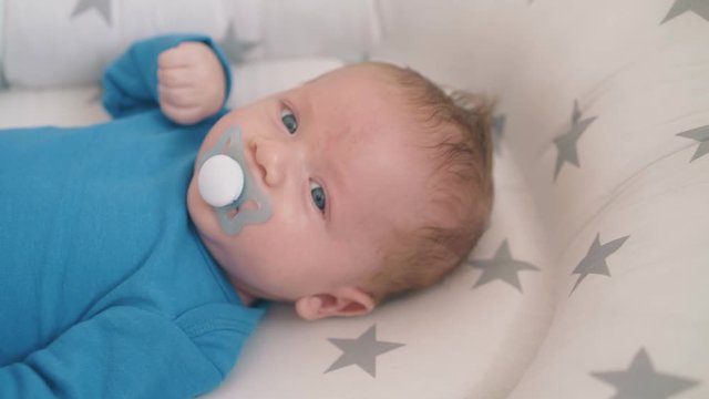 adorable little boy with bright blue eyes kicks legs sucking dummy in soft cocoon with stars in cot closeup