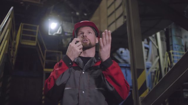 Waist-up shot of stressed Caucasian male technician on overalls and safety helmet standing on production line floor and talking on portable radio with frustrated and agitated expression