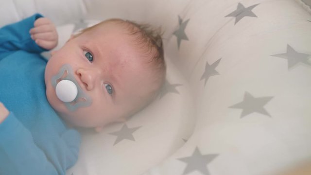 happy newborn baby sucks funny dummy lying in crib with comfortable cocoon in light room close view