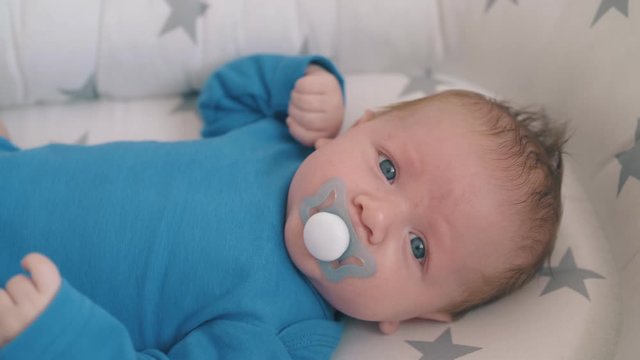 upset small baby sucks butterfly shaped dummy lying in cocoon in rocking crib at home closeup