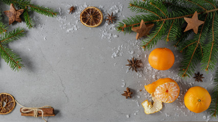 Christmas festive composition with three fresh mandarins, decorative stars on fir branches, anise, cinnamon and artificial snowflakes on a stone grey board. With space for text, top view