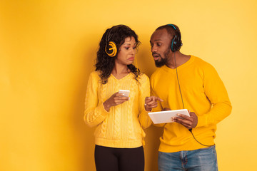 Young emotional african-american man and woman in bright casual clothes on yellow background. Beautiful couple. Concept of human emotions, facial expession, relations, ad. Using tablet and smartphone.