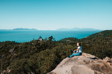 Young stylish independent girl travel the world, sitting on the rock and enjoying the view over the ocean and trees