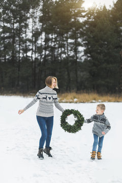 happy family mother and child son in jeans and knitted sweaters on a winter walk outdoors, running and having fun, holding home made Christmas wreath