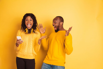 Young emotional african-american man and woman in bright casual clothes on yellow background. Beautiful couple. Concept of human emotions, facial expession, relations. Shocked pointing on smartphone.