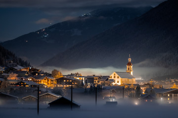 Winter night landscape overlooking the Austrian Tyrolean city of Neustift and the Pfarre church against the backdrop of mountains and clouds. Frosty night with fog in the valley