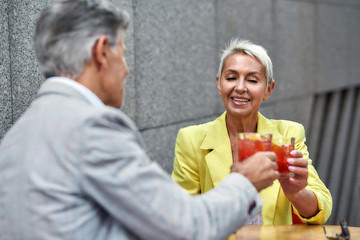 Cheers. Cheerful and beautiful middle-aged woman in yellow suit jacket drinking cocktails with her friend and smiling while sitting in cafe outdoors together