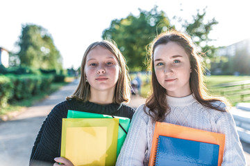 Two schoolgirls girlfriends, in summer in park are standing with notebooks notes. Happy smiles, in warm sweaters autumn day. concept learning at college school, outdoor recreation after class.