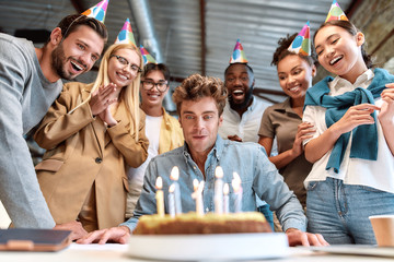 Corporate party. Young happy man blowing candles on cake while celebrating birthday with cheerful colleagues wearing party hats