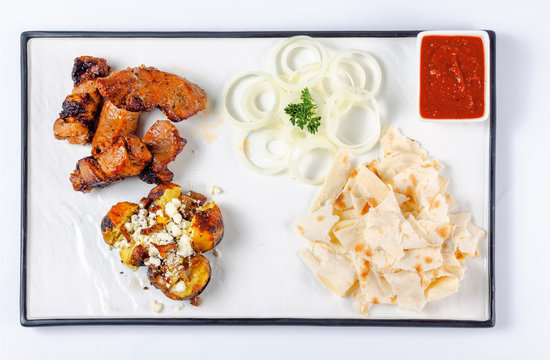 grilled pork and baked potato with cheese. onion circles and pita bread, red sauce on a white board. great snack dish for beer. top view, flat layout