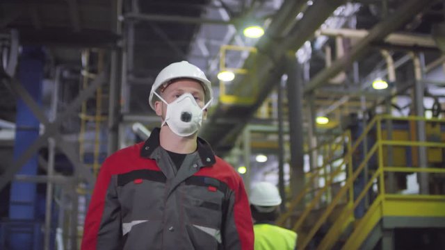Chest-up tracking shot of Caucasian male technician in uniform work clothes and hardhat putting protective dust mask on face and walking along mineral wool production line at manufacturing factory
