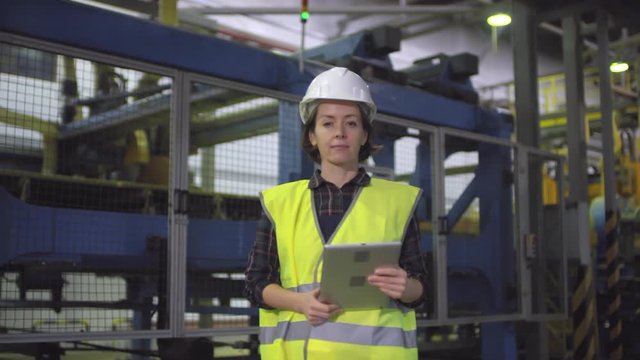 Waist-up arc shot of confident young Caucasian female operator standing in front of cutting machine on production line at mineral wool manufacturing plant, holding tablet and looking at camera