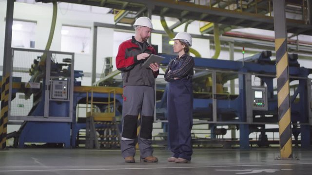 Full arc shot of Caucasian male and female manufacturing plant employees in hardhats standing in cavernous production hall in front of automated machinery, man explaining and woman listening