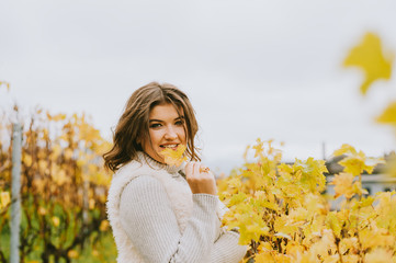 Autumn portrait of happy young woman enjoying nice day in vineyards, wearing grey pullover and fluffy white jacket, holding a leaf