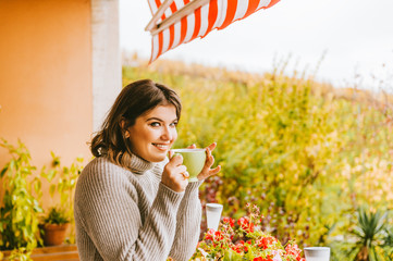 Romantic portrait of beautiful young woman wearing grey pullover, relaxing on the balcony between...