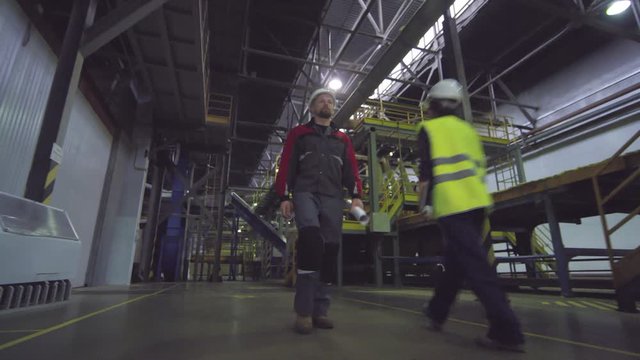 Low-angle full tracking shot of Caucasian male industrial factory worker in working uniform and safety helmet walking along rockwool production line at manufacturing plant, and colleague passing by