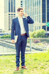 Young businessman traveling, working in New York City, wearing blue suit, white shirt, brown leather shoes, holding laptop computer, standing on green lawn in business district, talking on cell phone.