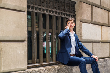 Young man traveling in New York City, wearing blue suit, white shirt, sitting on big window frame of old street, talking on cell phone, looking, waiting, taking work break. Concept for city life..