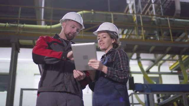 Low angle thigh-up arc shot of Caucasian man and woman in work uniform and safety helmets standing together in production hall of manufacturing plant, using tablet and discussing technological issues
