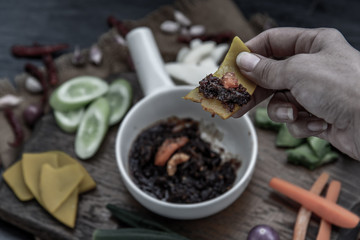 The hand is using pumpkin to scoop fried chili paste with shrimps (thai language nam prik pao) with the ingredient and fresh vegetables on wood background.