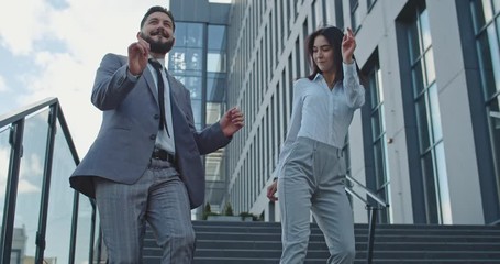 Happy and joyful Caucasian young businessman and businesswoman walking down the steps while leaving office center building and dancing as got new job of his dreams. Outdoors.