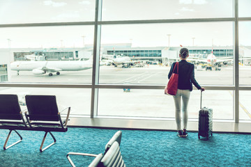 Traveler businesswoman waiting for delayed flight at airport lounge standing with luggage watching tarmac at airport window. Woman at boarding gate before departure. Travel lifestyle.