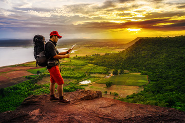 man traveler with backpack looking at the map on edge of cliff, on a top of the rock mountain at sunset