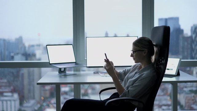 Female economist in classic glasses making research in social medial checking news and online content on smartphone gadget during time for working with market at office interior, corporate company