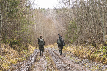 two men, in camouflage, hunters, are walking along a dirty road, looking for prey, with weapons