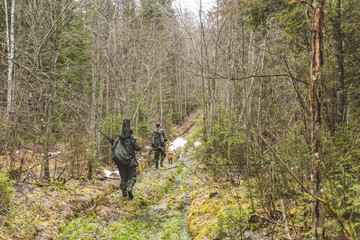 two men, in camouflage, hunters, are walking along a dirty road, looking for prey, with weapons