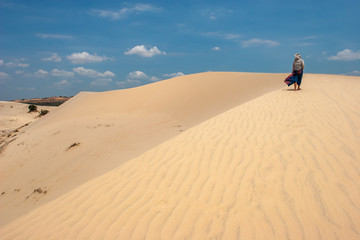 A girl walks along a large sand dune in the wind. Blue skirt flutters. Hood on the head.