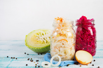 Two glass jars of fermented cabbage are on the table, around fresh vegetables, cabbage and onions. Copy space