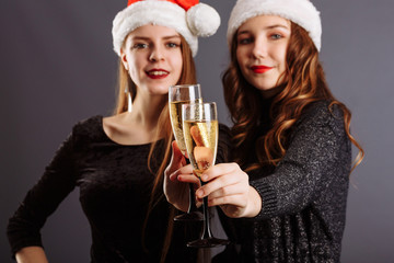 Two attractive cheerful girls with curly long hair in Christmas hat smiling, holding a champagne, posing to the camera, isolated on gray background