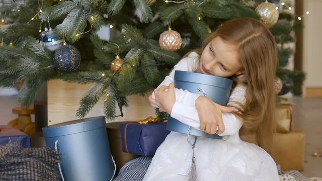 Smiling cute blond girl is hugging a blue gift box she recieved sitting near christmas tree, happy childhood and xmas celebration concepts