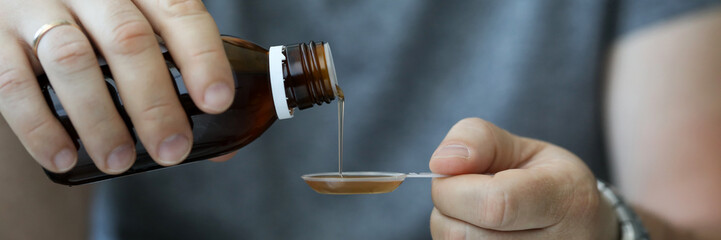 Focus on male hands holding bottle of cough syrup and measuring spoon. Guy measures desired dosage of medicine. Healthcare and treatment concept. Blurred background
