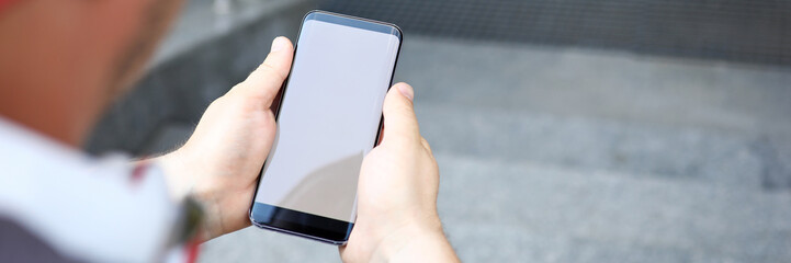 Focus on male hands holding modern cellphone. Man sitting on stairs with sensory mobile phone and messaging with friends. Copy space on screen of gadget. Blurred background