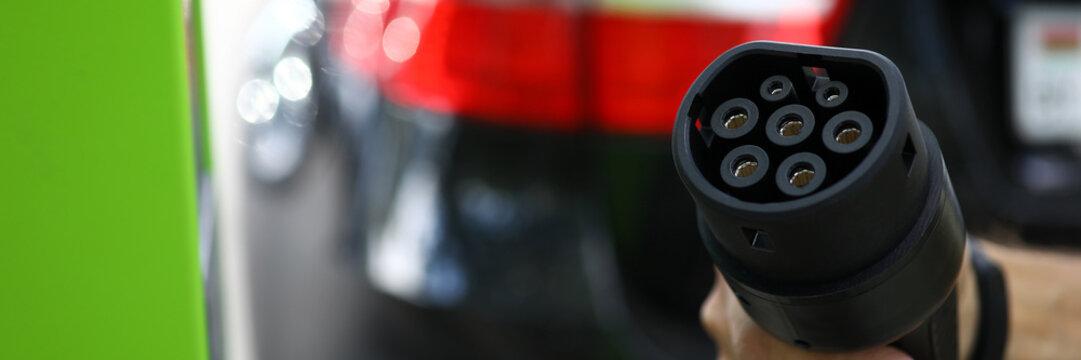 Focus on male hand holding modern charge device for cars. Automobile at filling station for electrocars. Environmental protection and newest transport technology concept. Blurred background